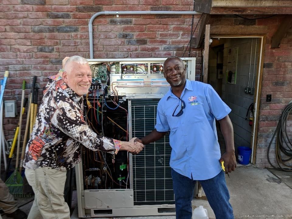 Two men posing beside a large metal HVAC unit in front of a brick building