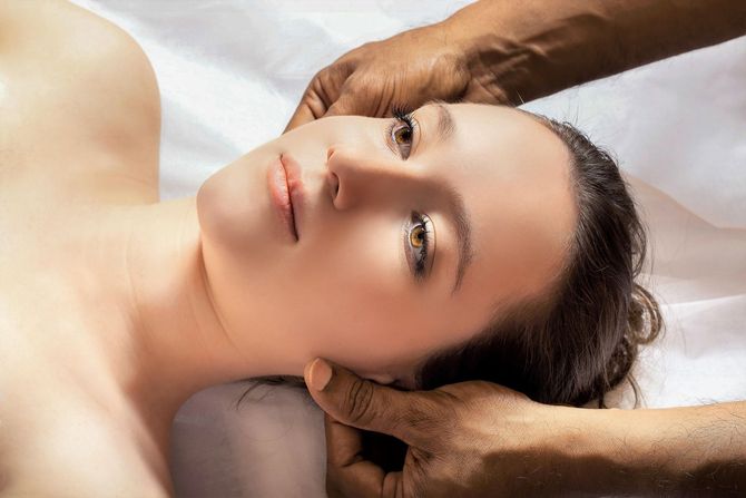 A person lies on a white surface receiving a relaxing facial or head massage from a pair of hands.