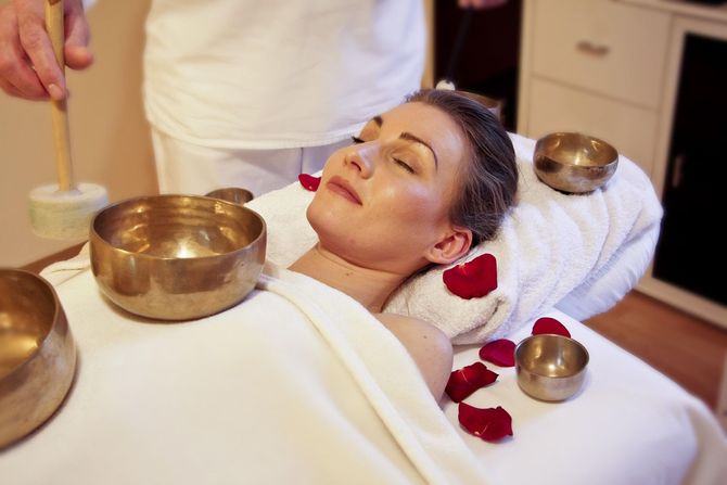 A person with closed eyes lies on a massage table during a sound therapy session with singing bowls and rose petals.