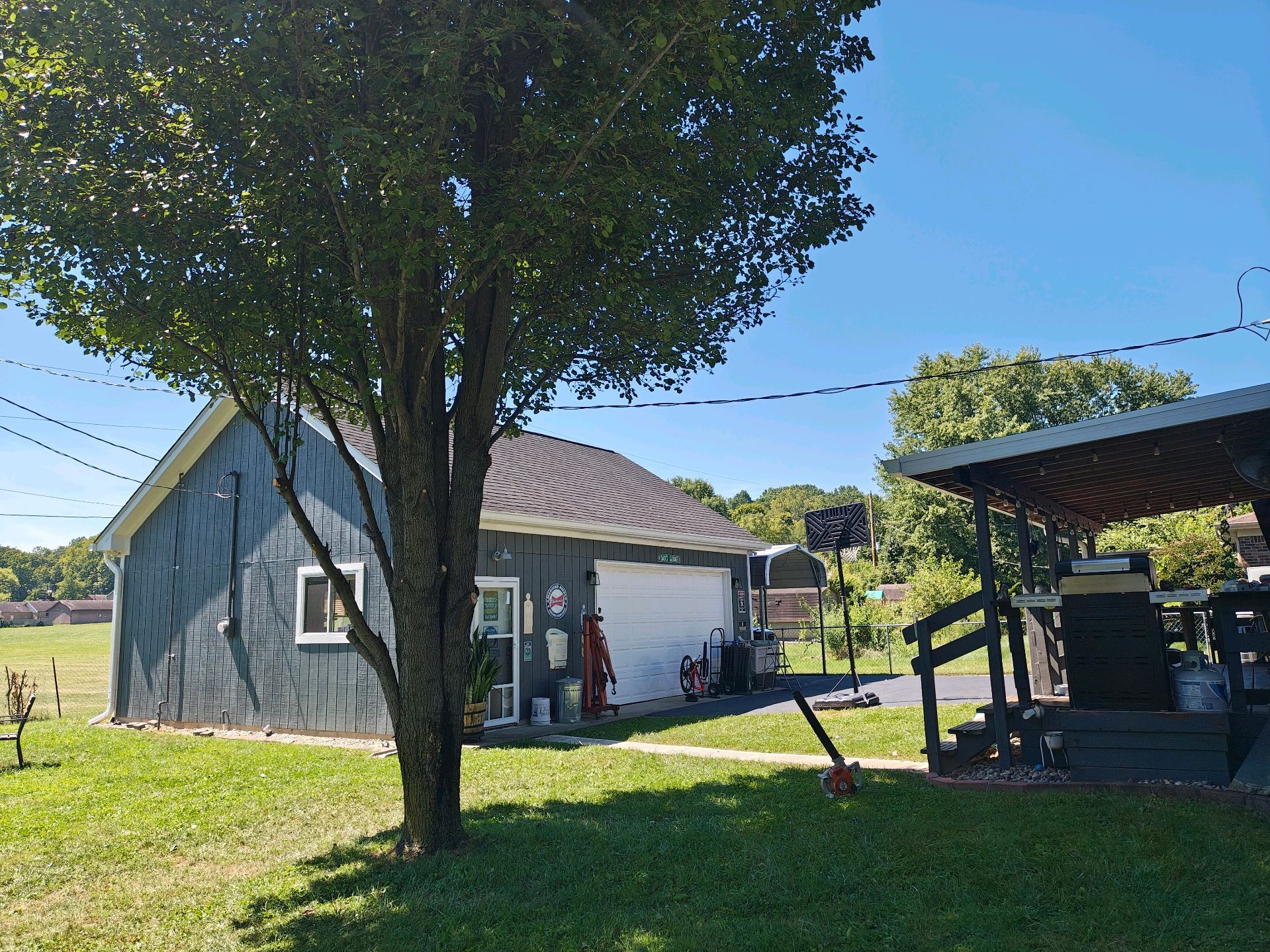 Gray shed and carport with tree in front on a sunny day.