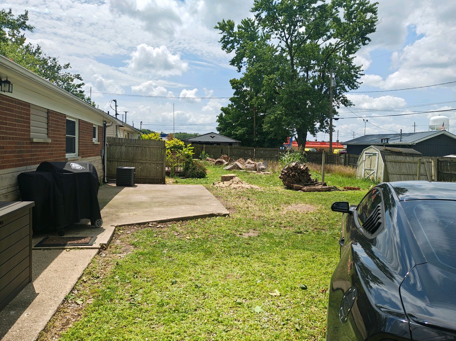 Backyard view, a brick house on the left, a car on the right, with overgrown grass, a concrete slab, and trees.