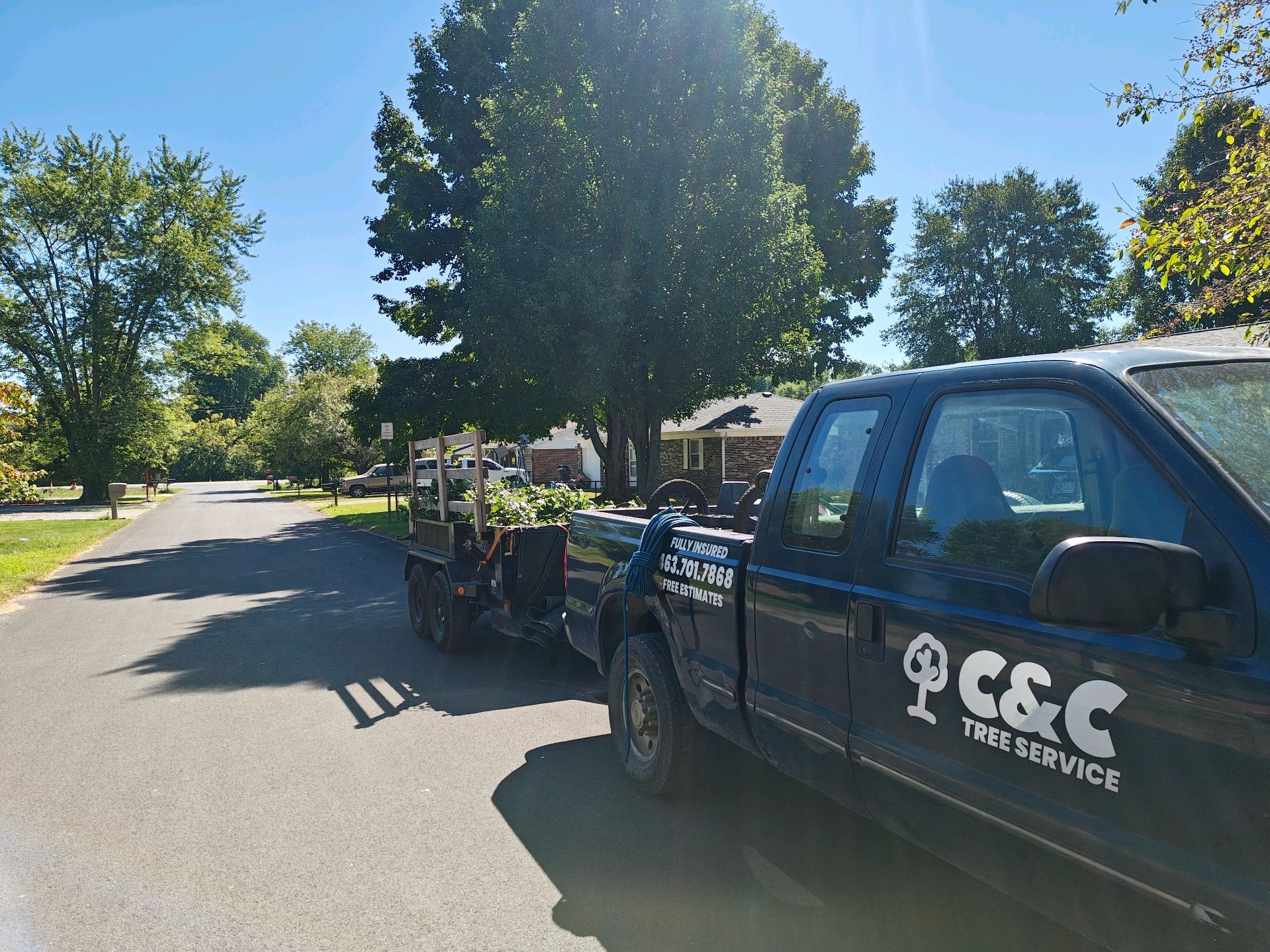 Truck from C&C Tree Service with a trailer loaded with branches parked on a residential street on a sunny day.