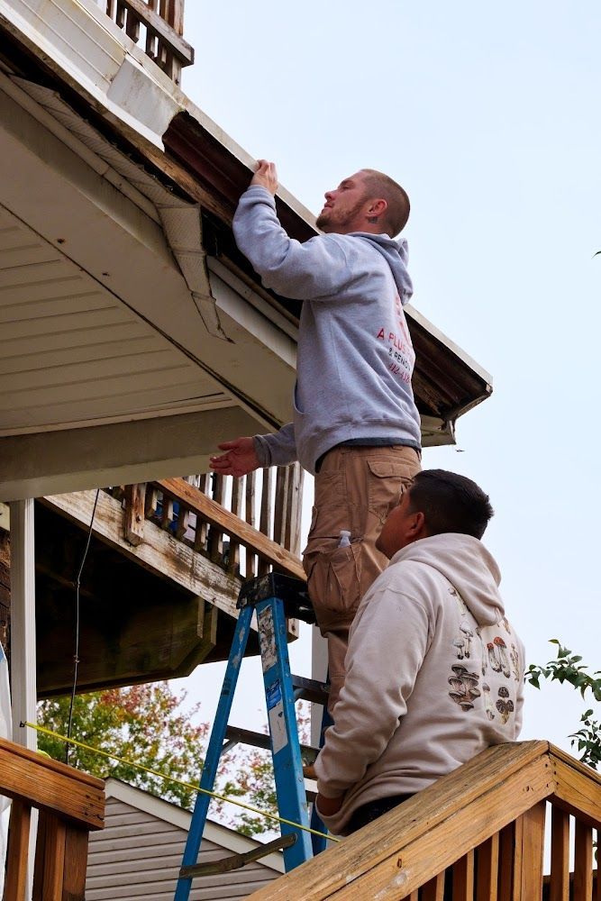 Two people on a ladder repairing a house's gutter. One person is at the top, reaching. Bright sky.