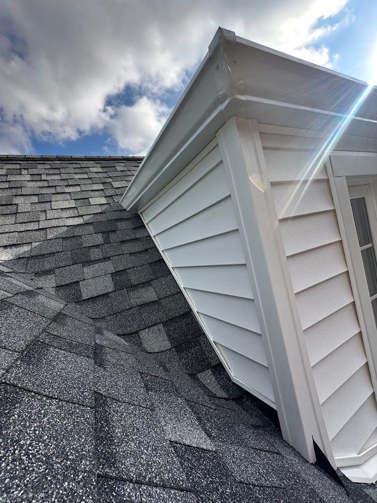 Roof with dark gray shingles, white siding on a dormer, against a cloudy sky.