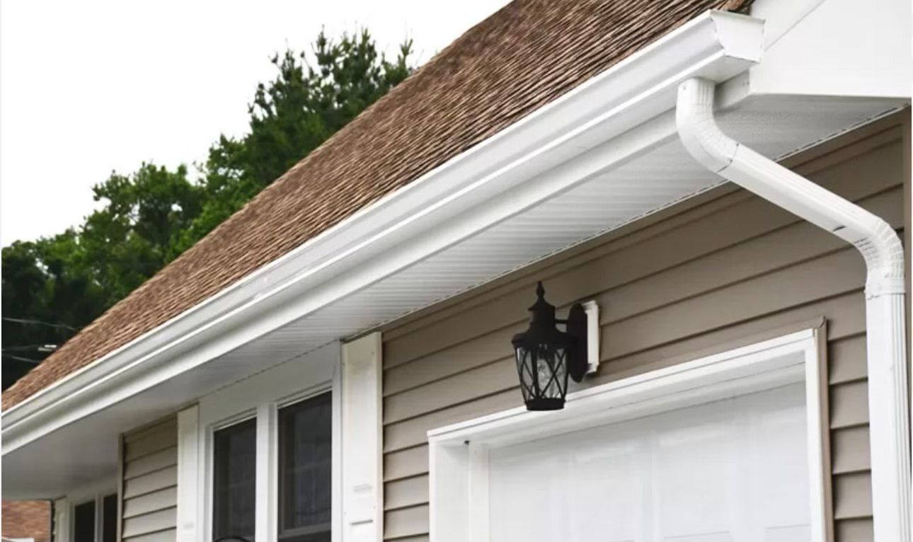 White gutters and downspout on a tan house with a brown shingled roof, trees in the background.