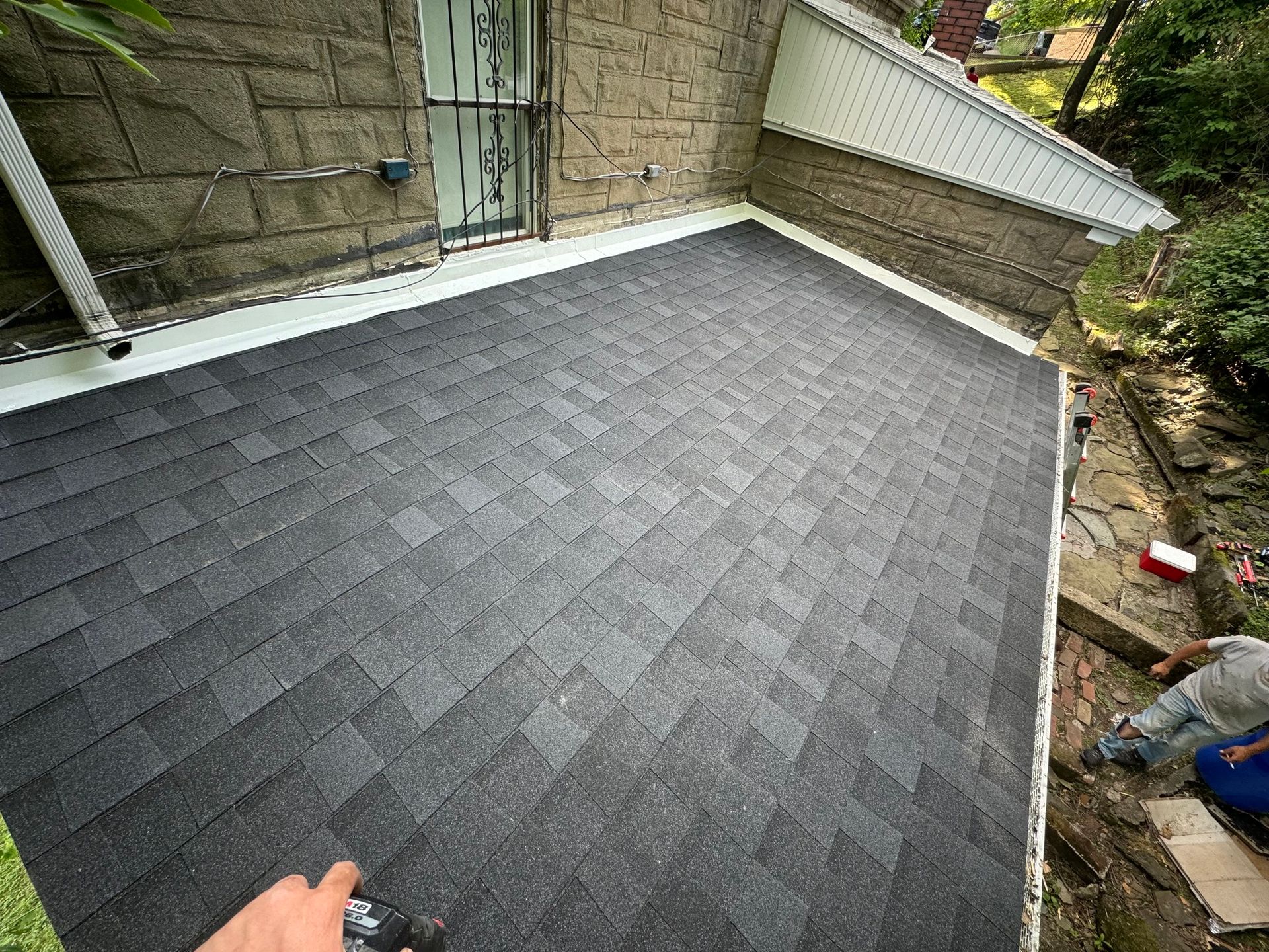 New asphalt shingle roof on a house, angled view. A person works near a gutter, trees visible in the background.