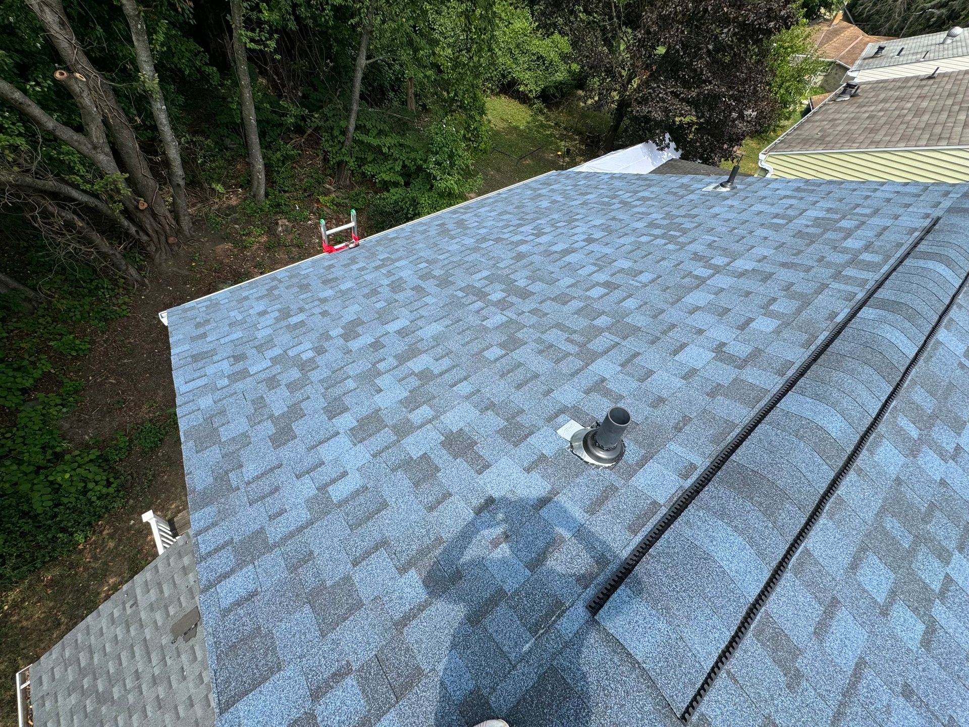 Blue asphalt shingle roof on a house, with a vent pipe and nearby trees.