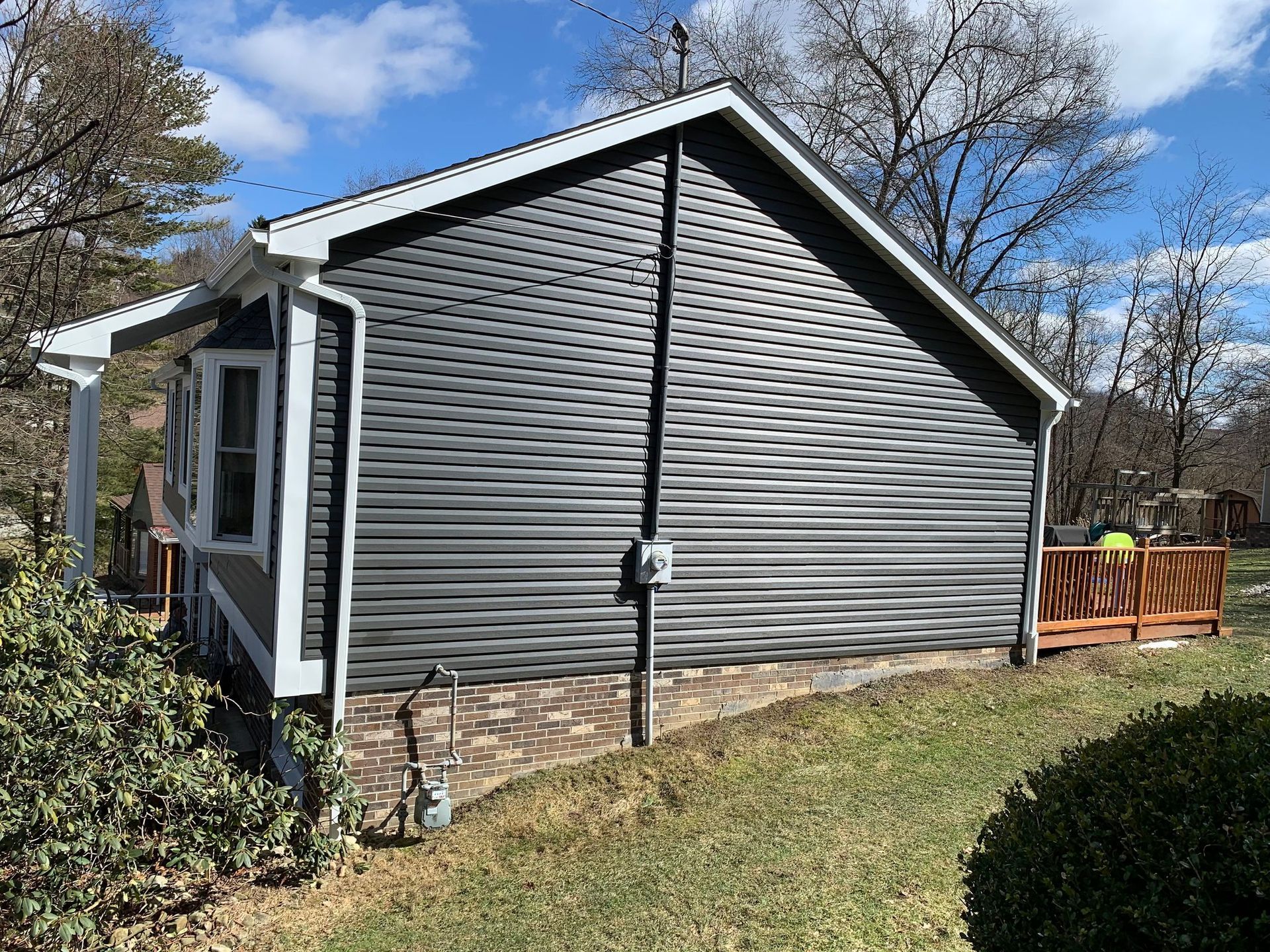 Side view of a house with dark gray siding, a brick foundation, and a small wooden deck.