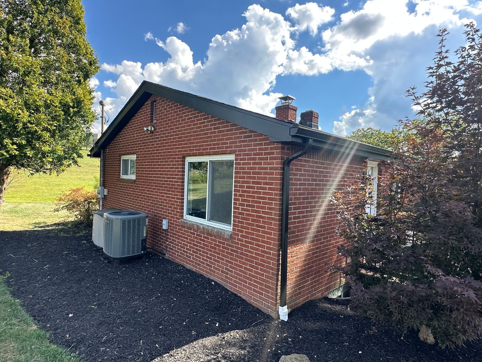 Brick house with black trim, surrounded by mulch, green grass, and trees under a cloudy blue sky.