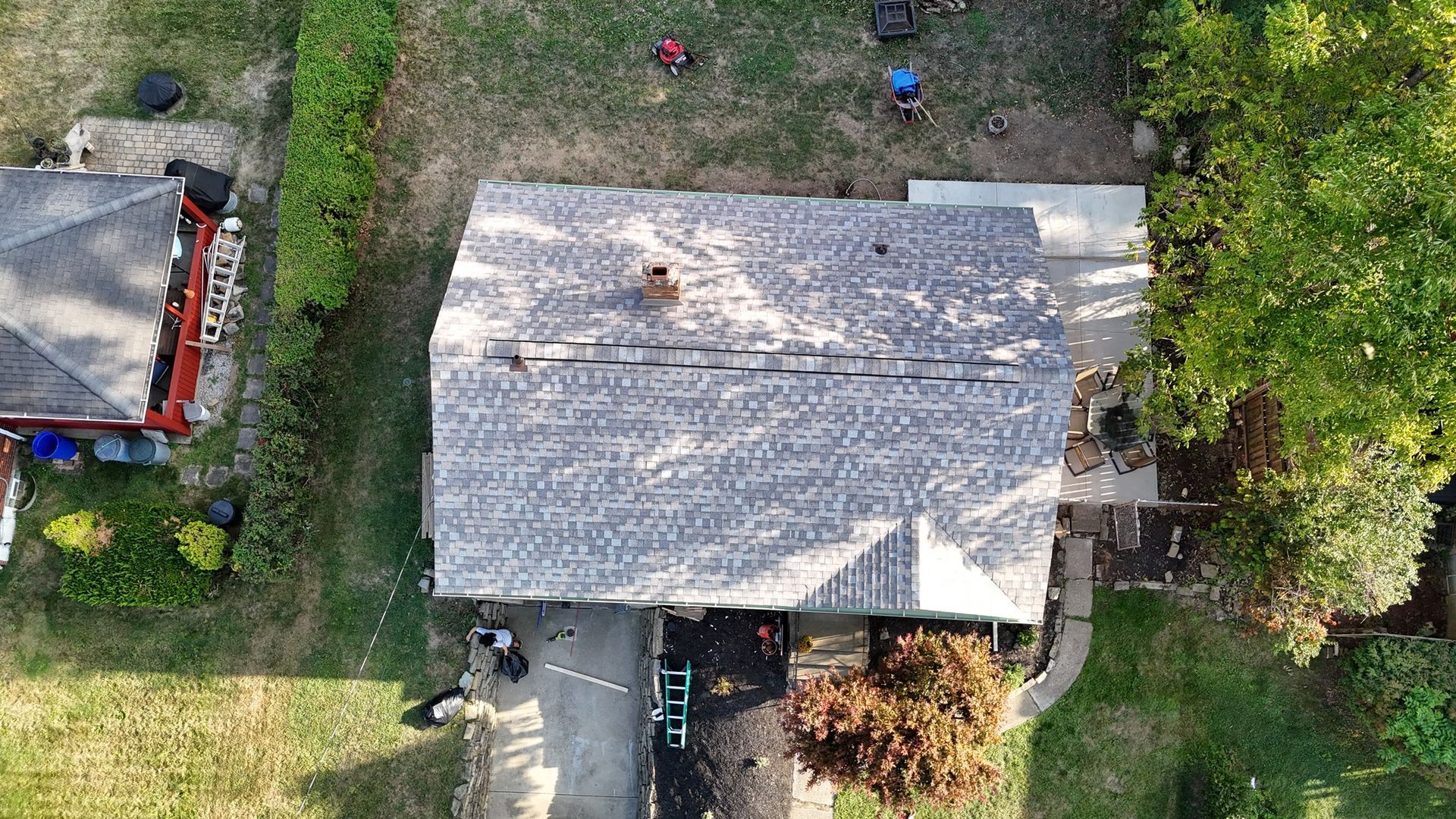 Aerial view of a house with roofing work in progress. Roof is gray with several workers. Green lawn and trees surround.