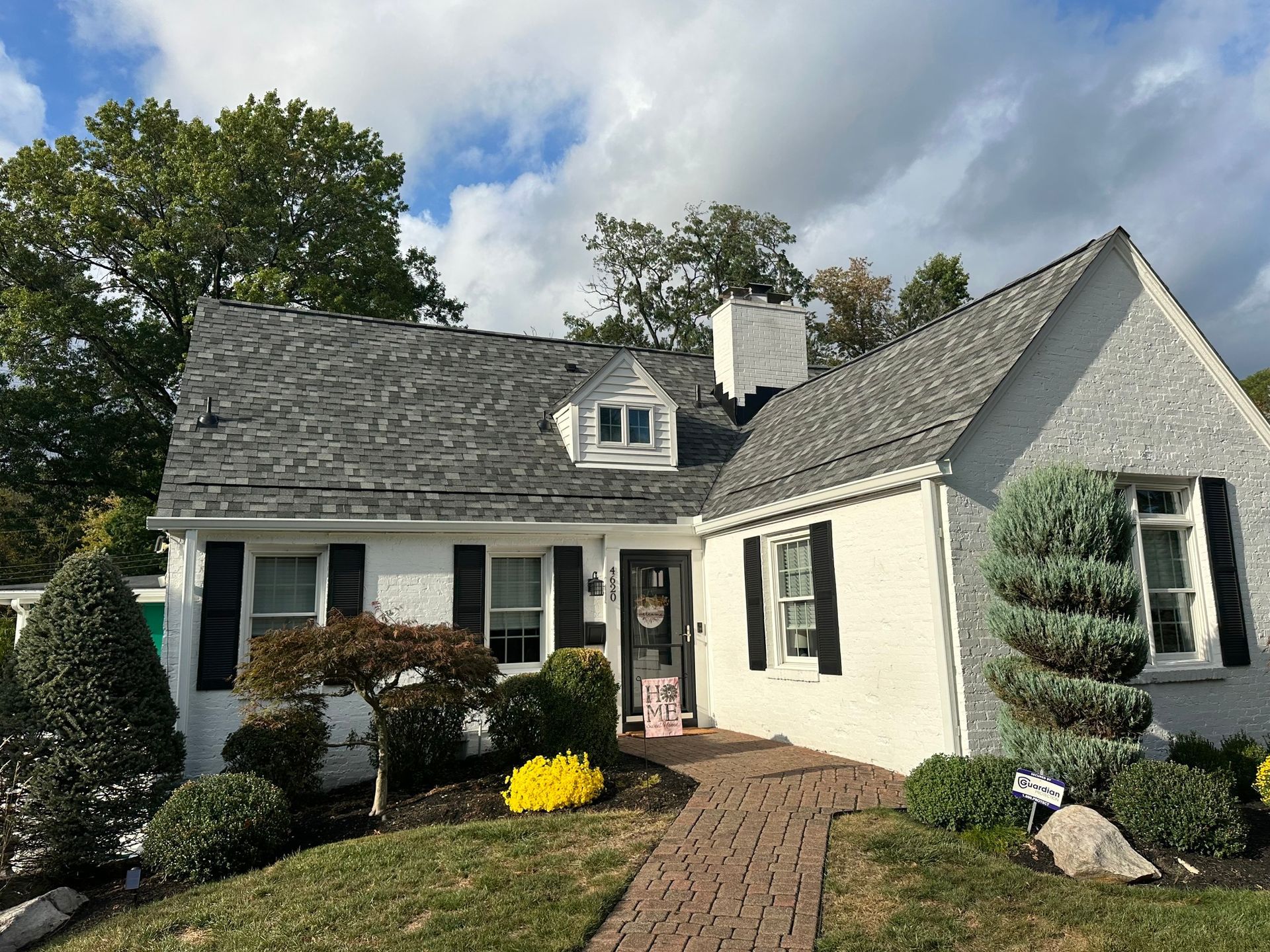 White house with gray roof, black shutters, brick path, and landscaped yard under a cloudy sky.