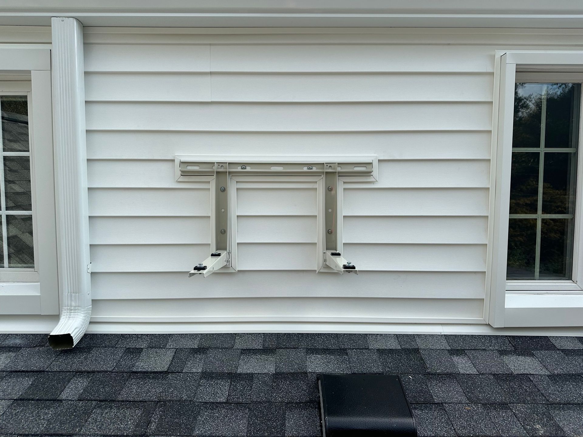 White siding with an empty bracket for an AC unit, between two windows and above a roof.