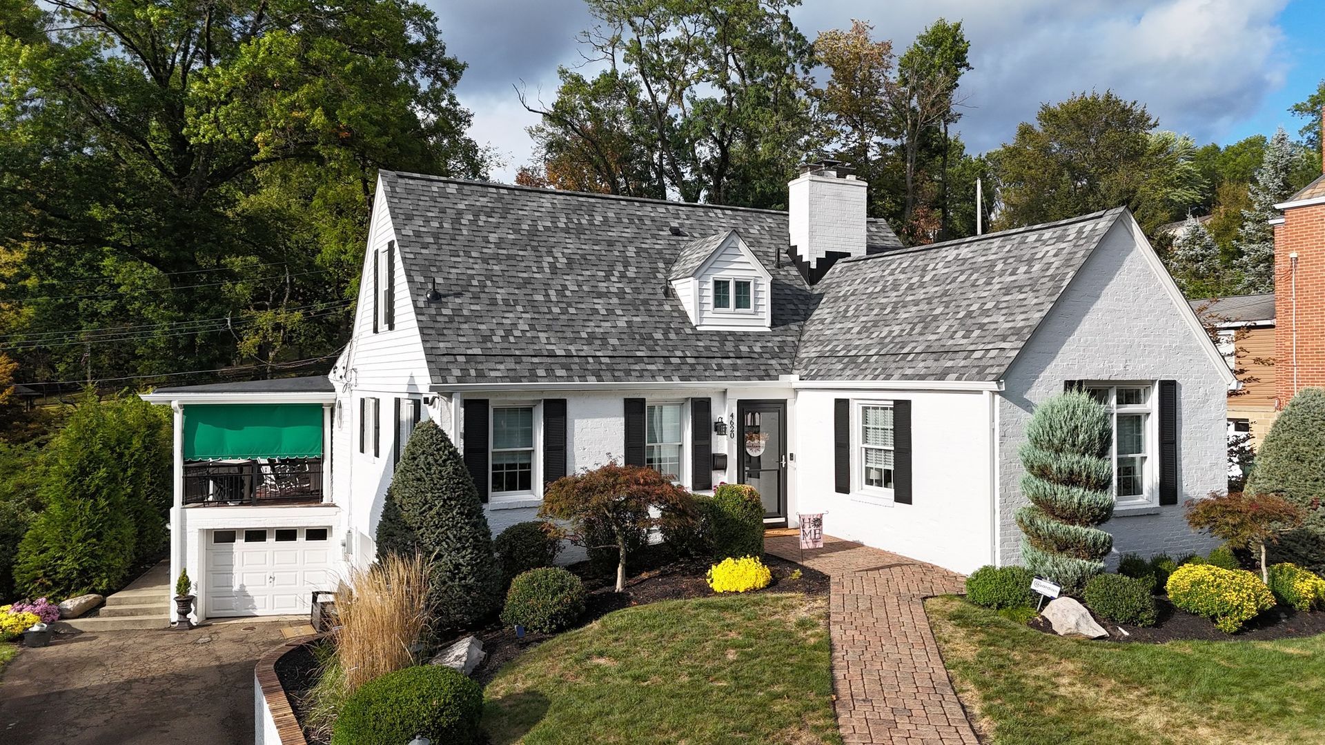 White stucco house with dark shutters, gray roof, and brick walkway. Lush landscaping with green foliage.