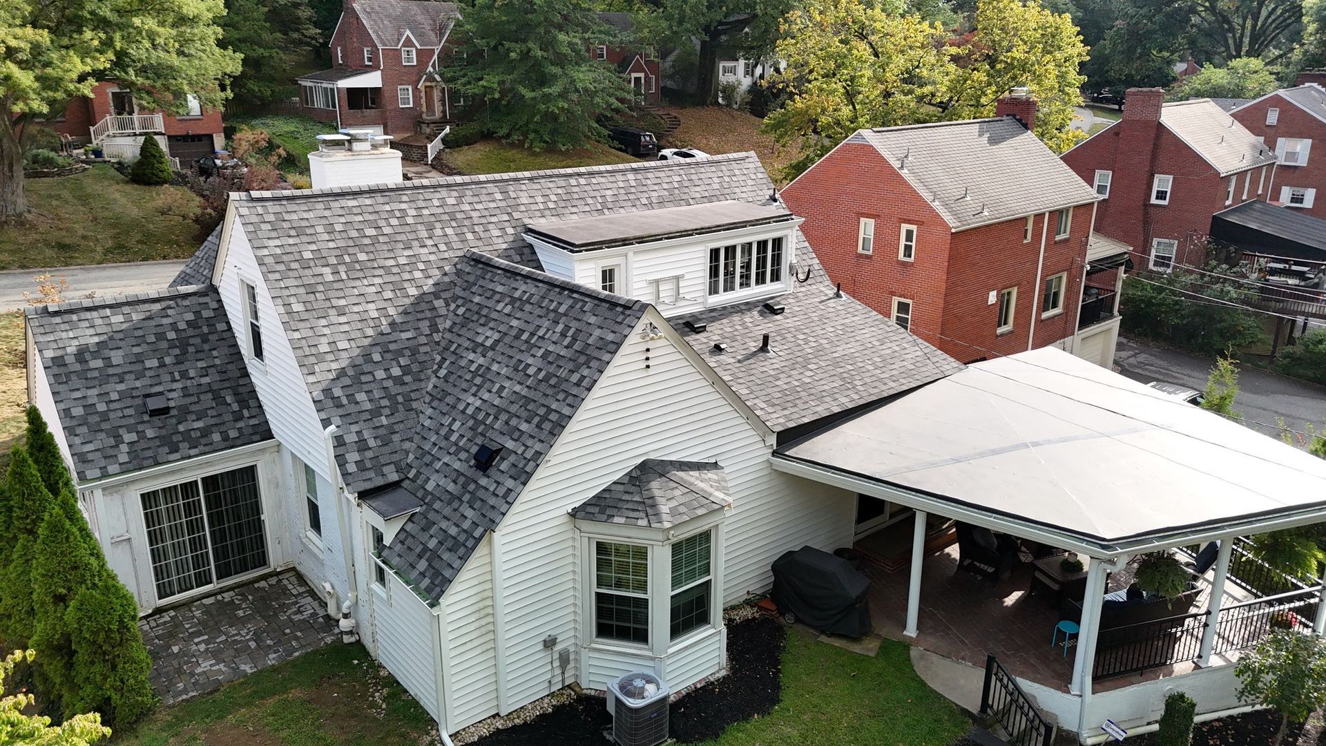 White house with gray shingle roof, attached porch, and red brick building in the background.