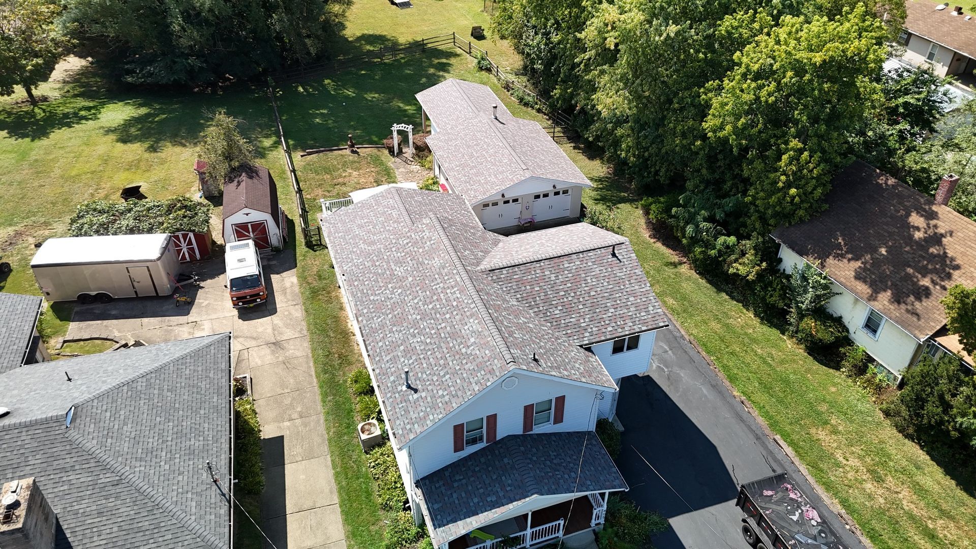 Aerial view of a two-story house with a long driveway. Brown and grey roof, green lawn, trees.