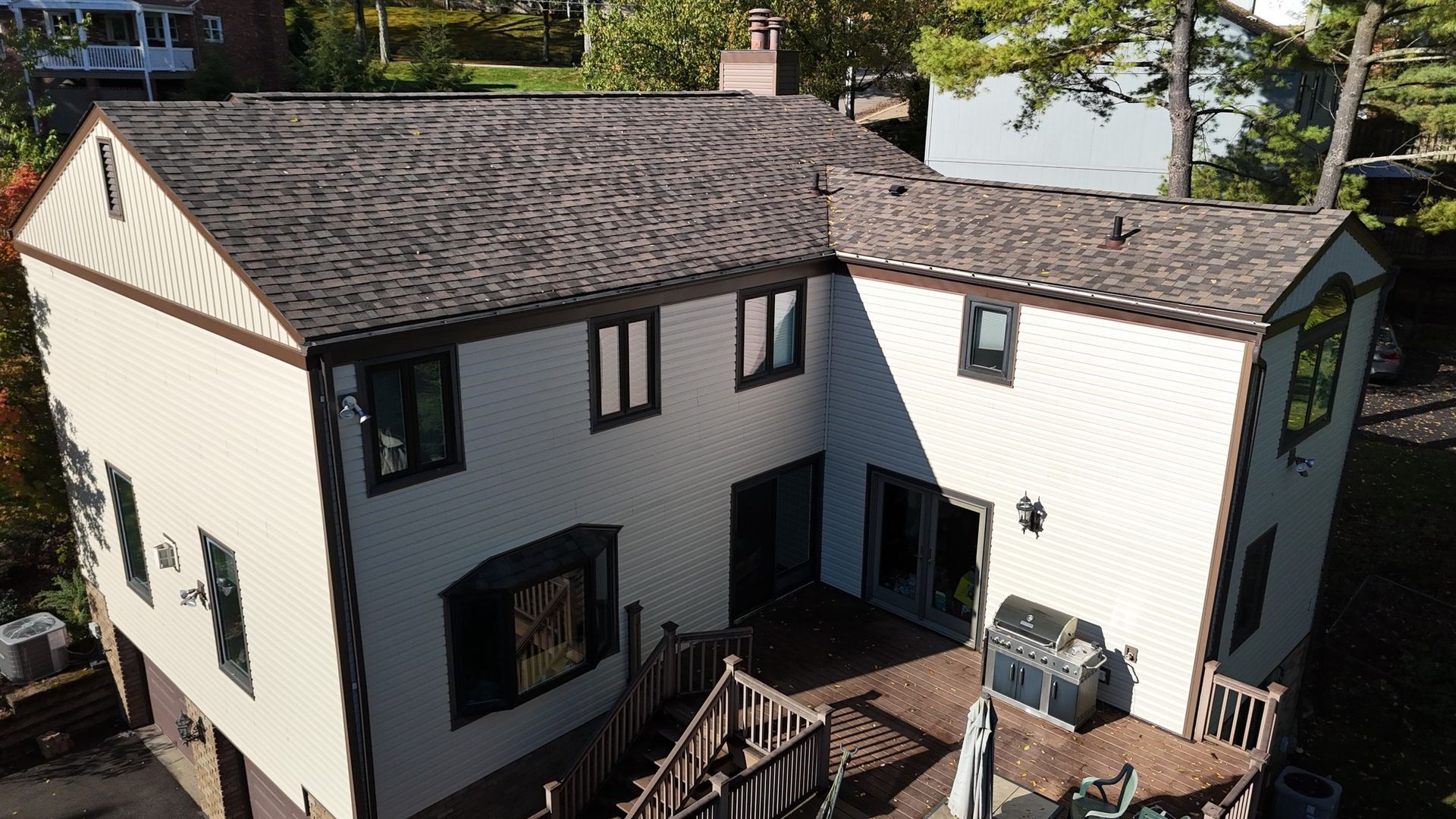 Two-story beige house with brown roof and trim, deck with grill, surrounded by trees.