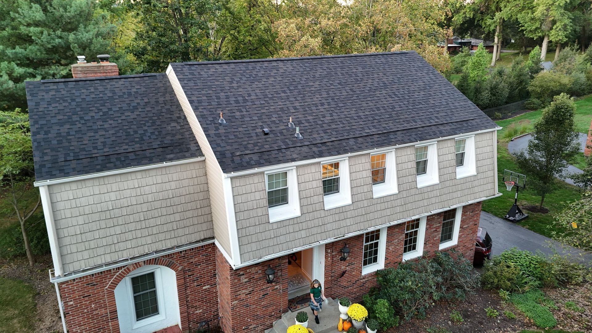 House with dark gray roof, red brick base, light brown siding, white trim, and trees in background.