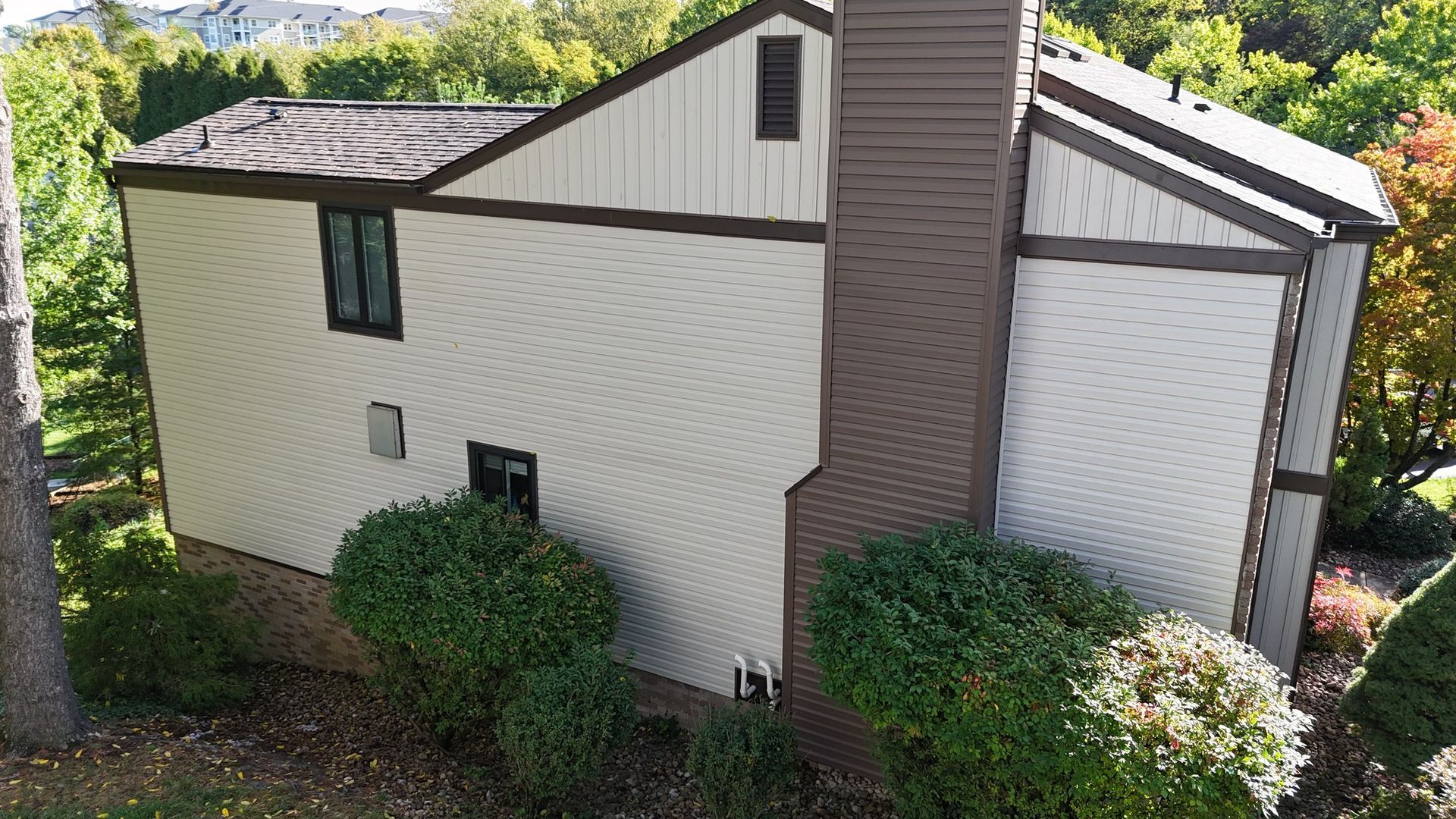 Beige sided house with brown trim and chimney, two visible windows, surrounded by trees.