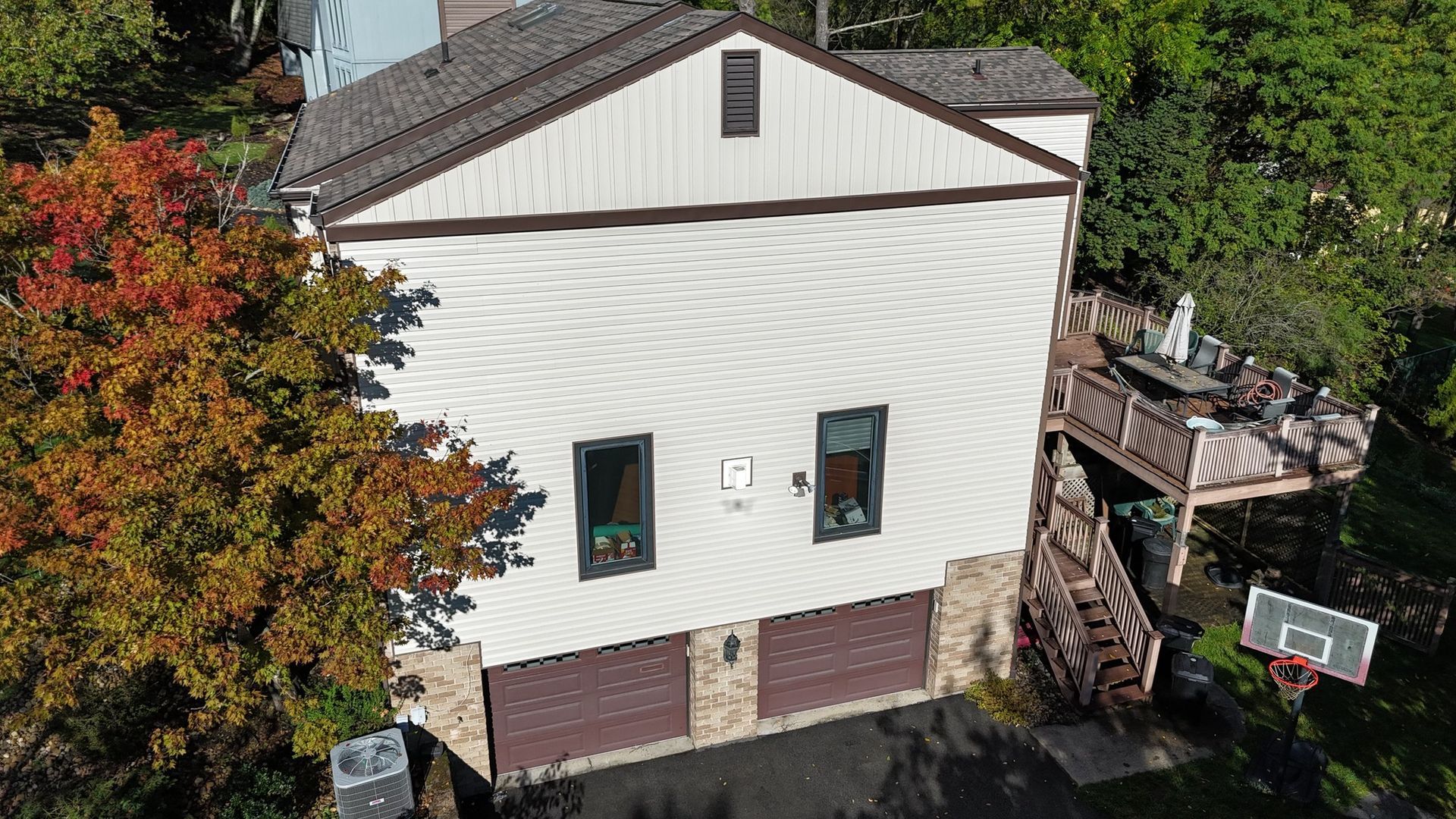Two-story house with brown garage doors, white siding, and a deck with a basketball hoop in yard.