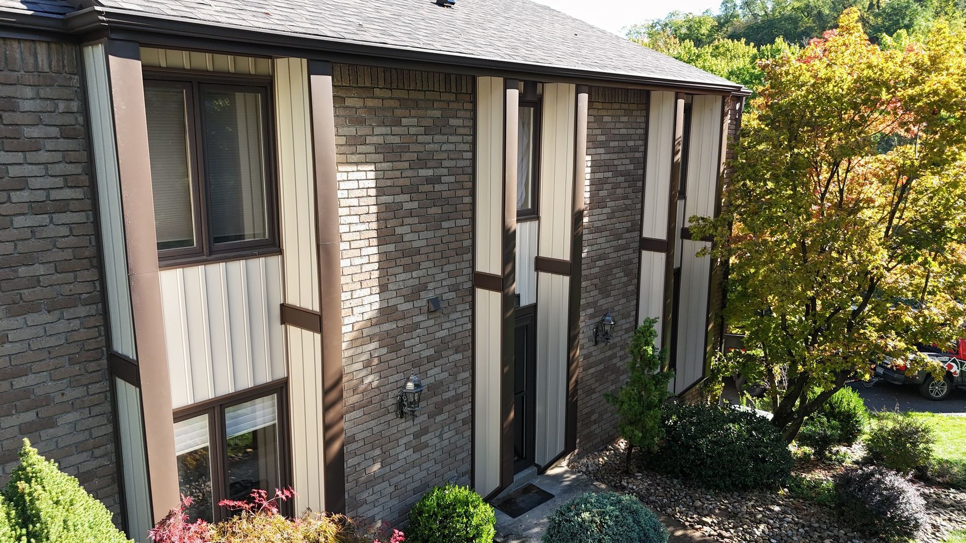 Brick house with brown trim and windows, surrounded by green shrubs and a tree.