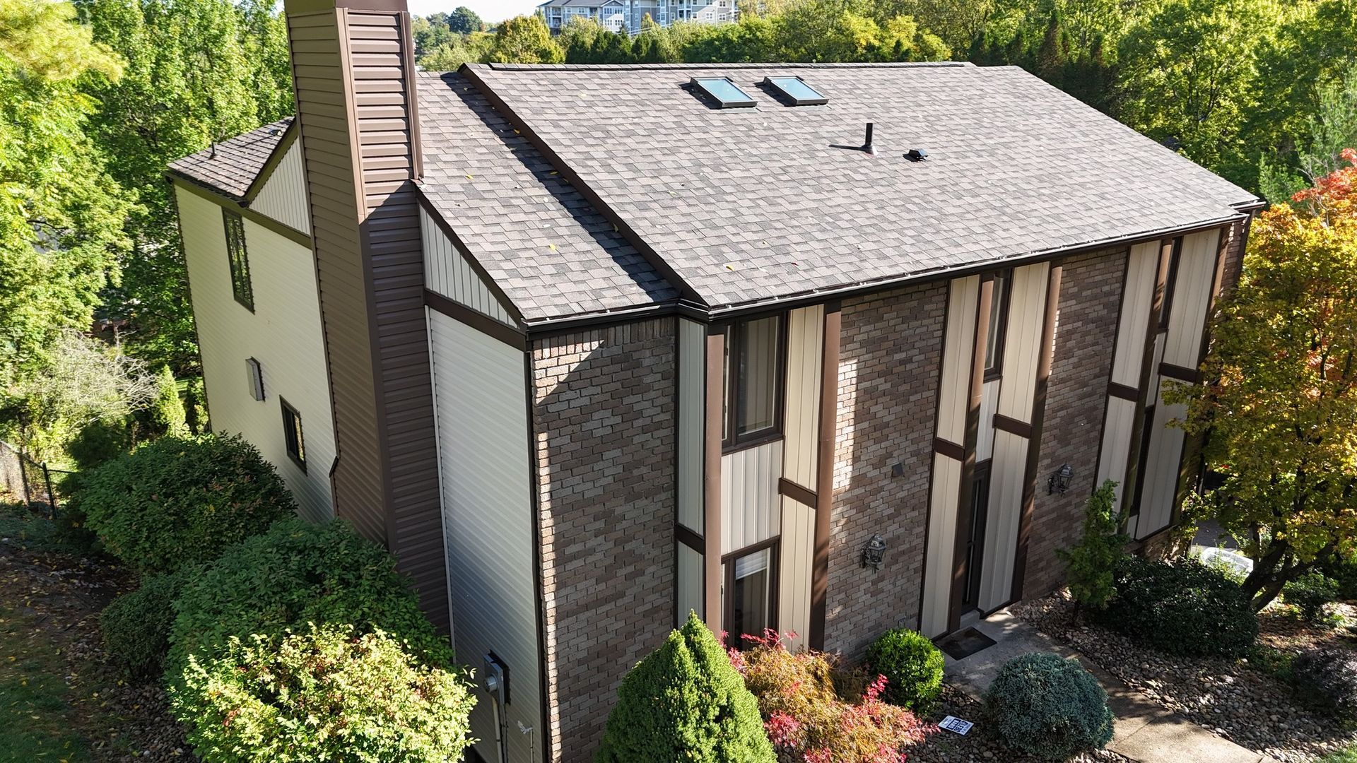 Two-story house with brown brick, tan siding, brown roof, and chimney, surrounded by trees.