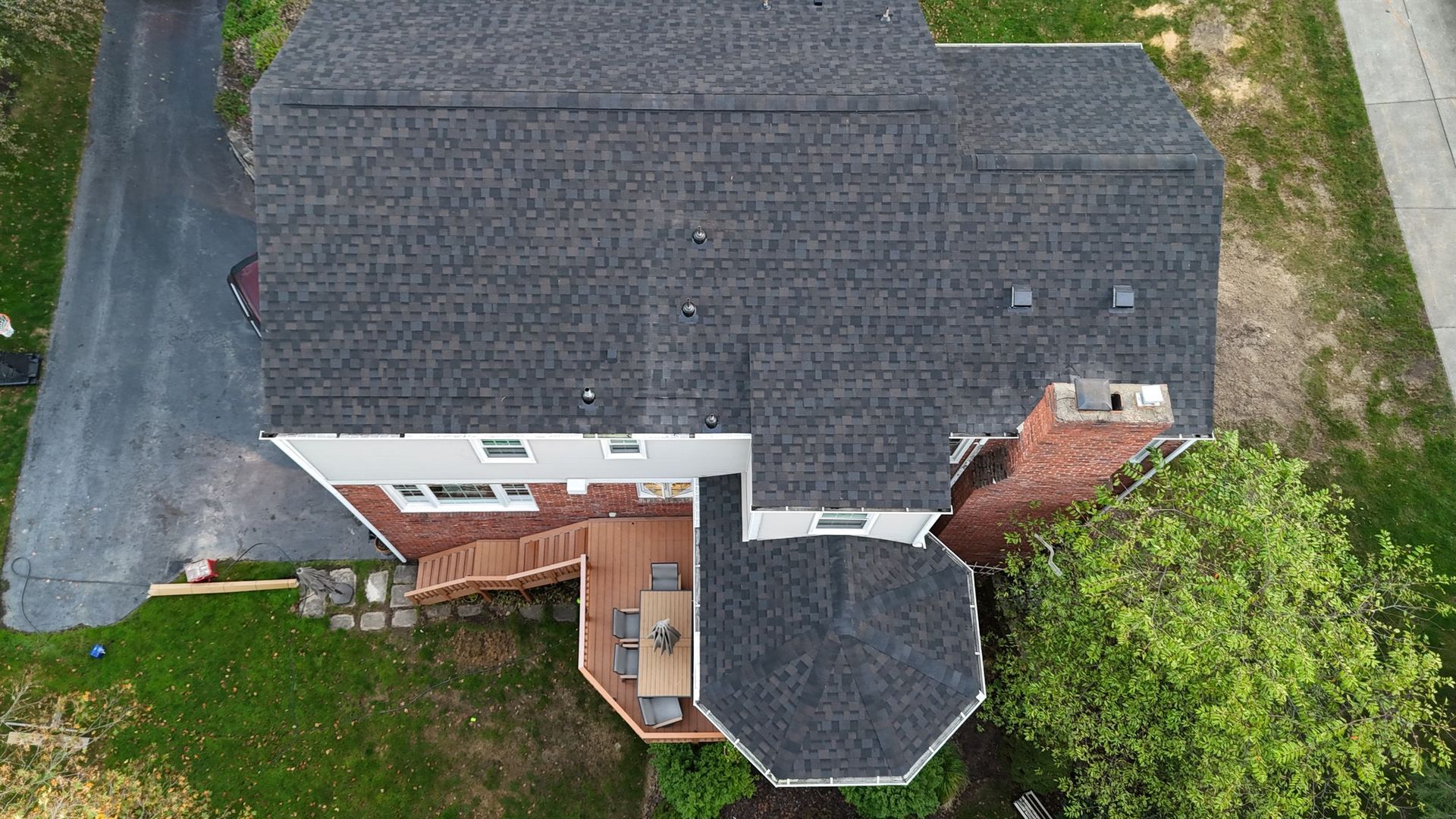 Overhead view of a house with a dark gray roof, red brick chimney, and wooden deck surrounded by green grass.