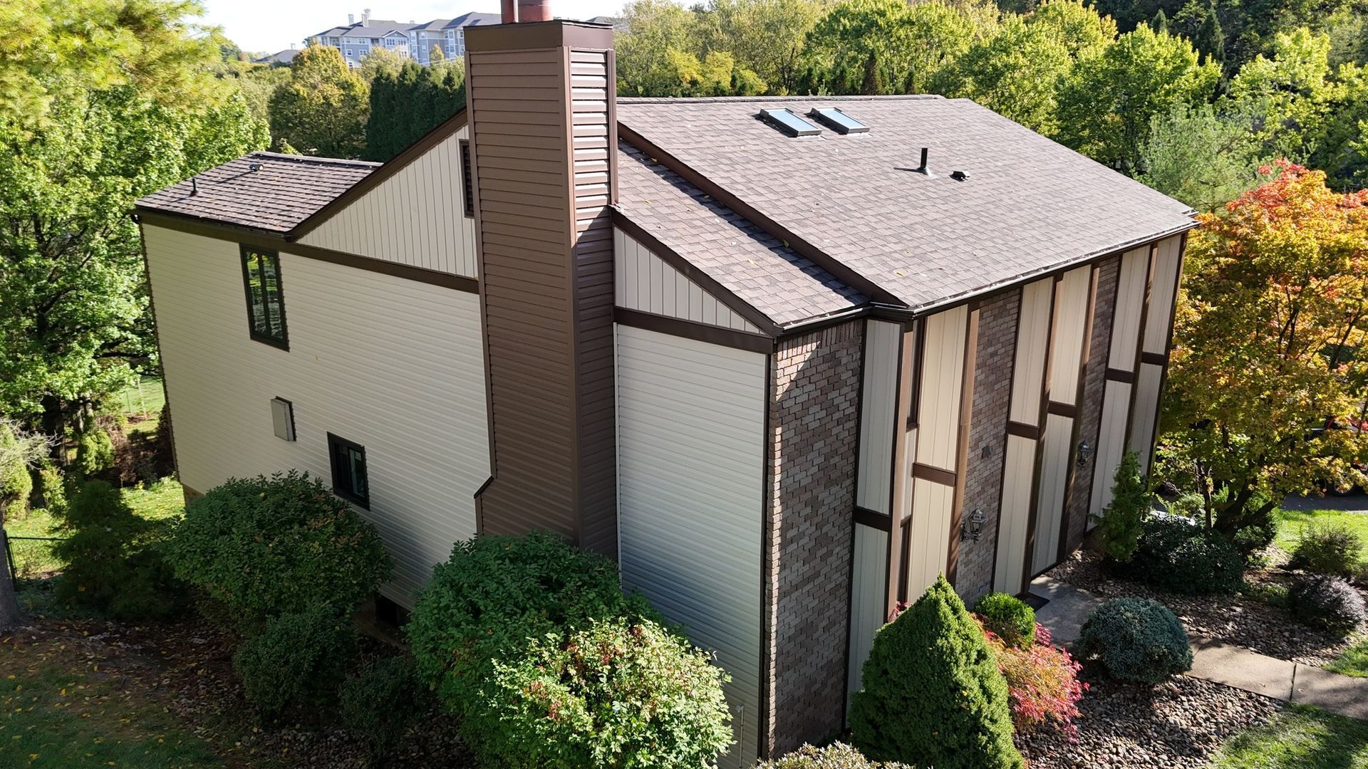 Two-story house with brown trim and roof, brick chimney, surrounded by green and autumn trees.