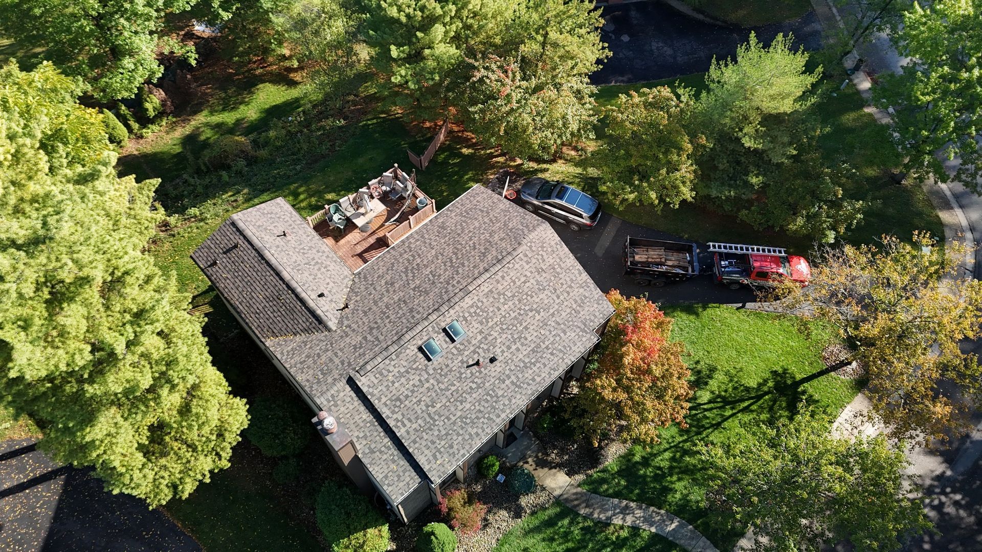Overhead view of a house with a gray roof, surrounded by trees and a driveway with vehicles.