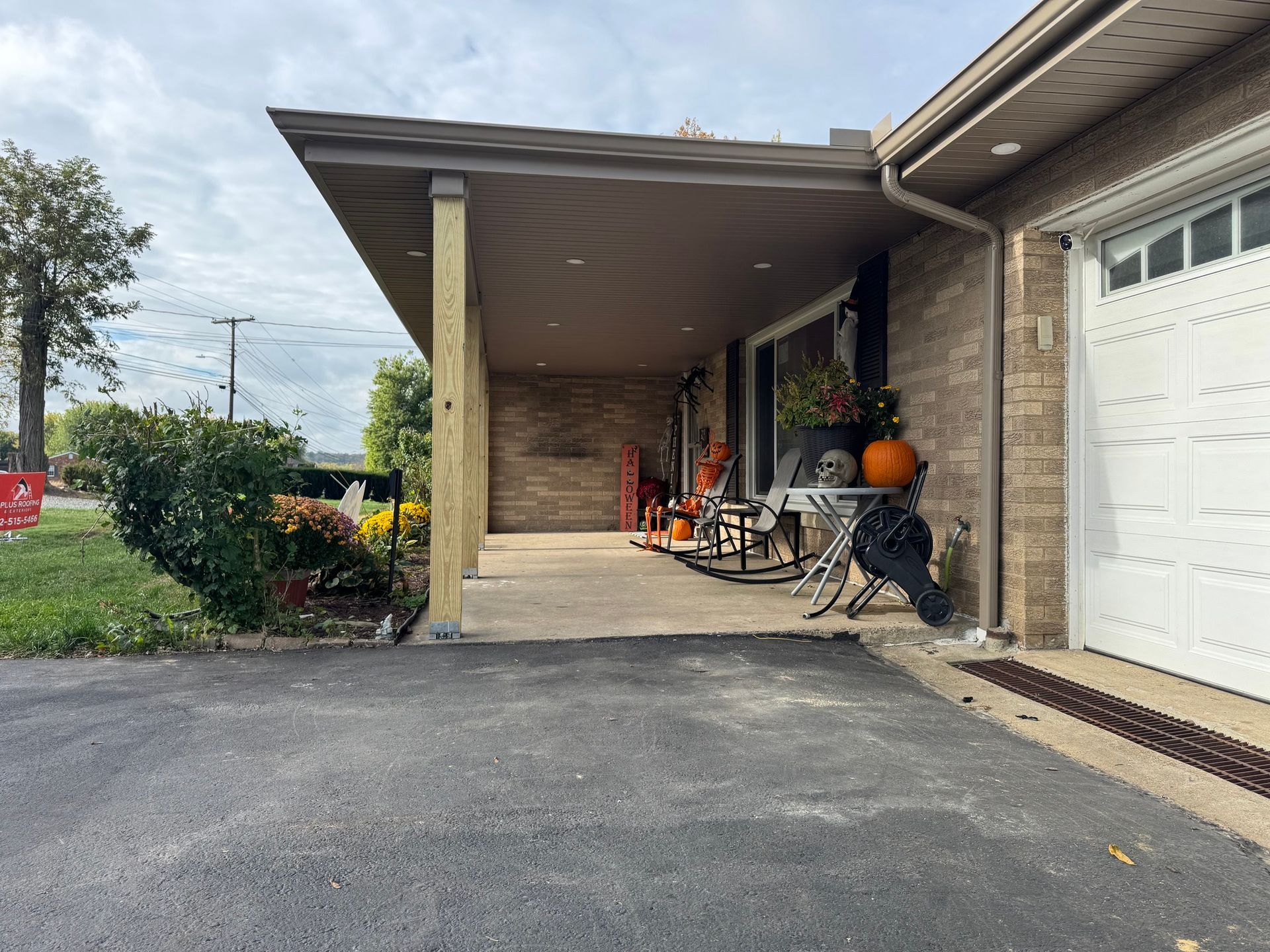 Covered porch with chairs, pumpkins, and a garage. Paved driveway, green shrubs, and a cloudy sky.