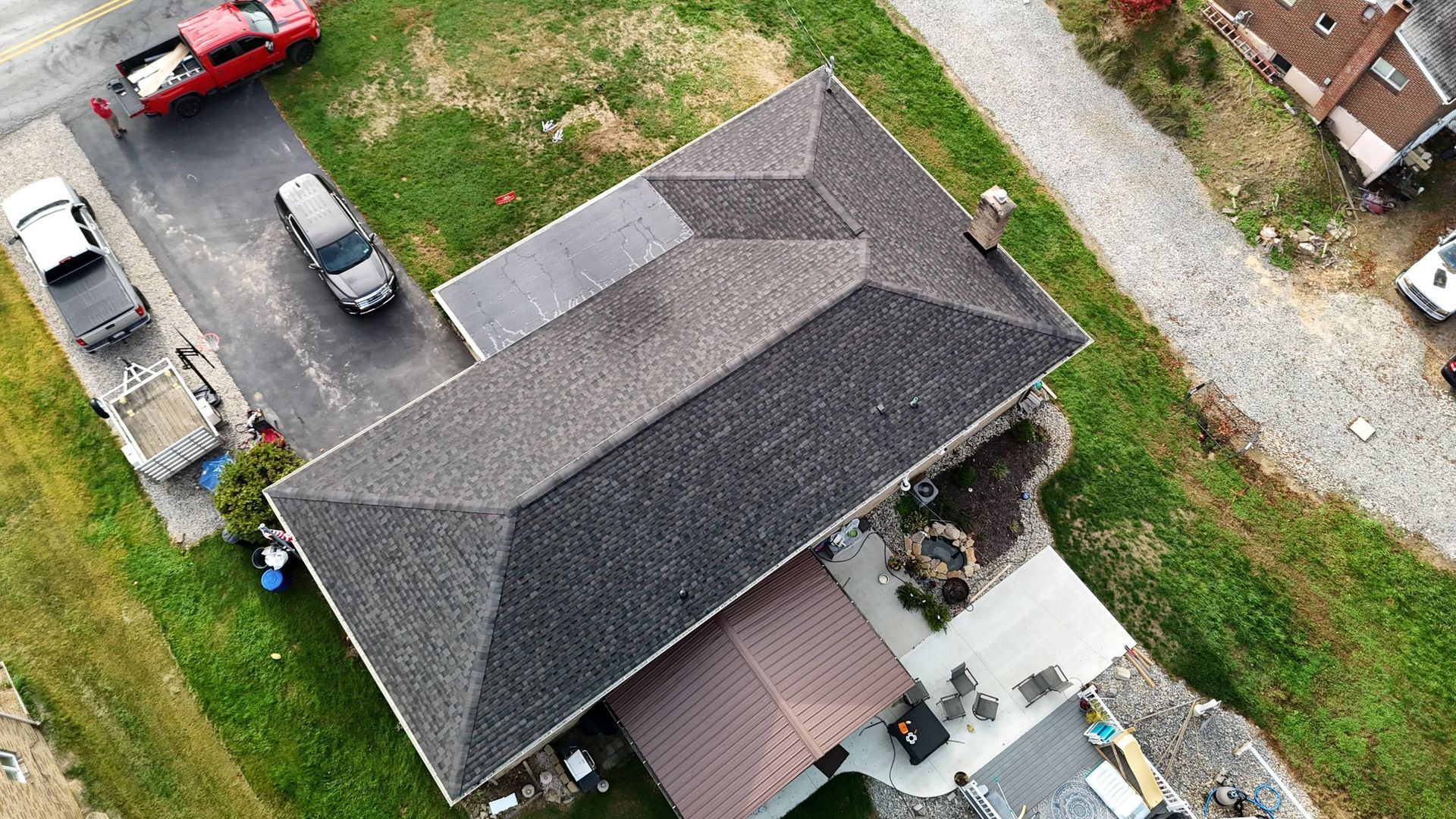 Overhead view of a house with a dark gray shingled roof, a brown patio roof, and a driveway with vehicles.