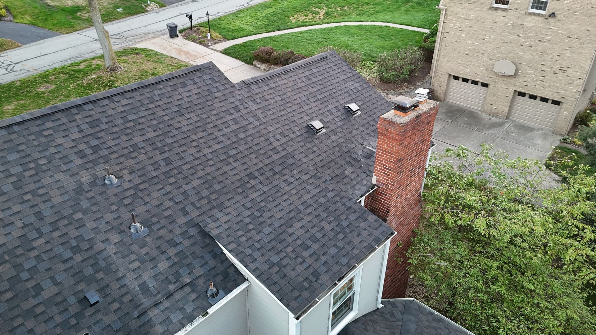 Overhead view of a house with a dark gray shingle roof, brick chimney, and a neighboring building.