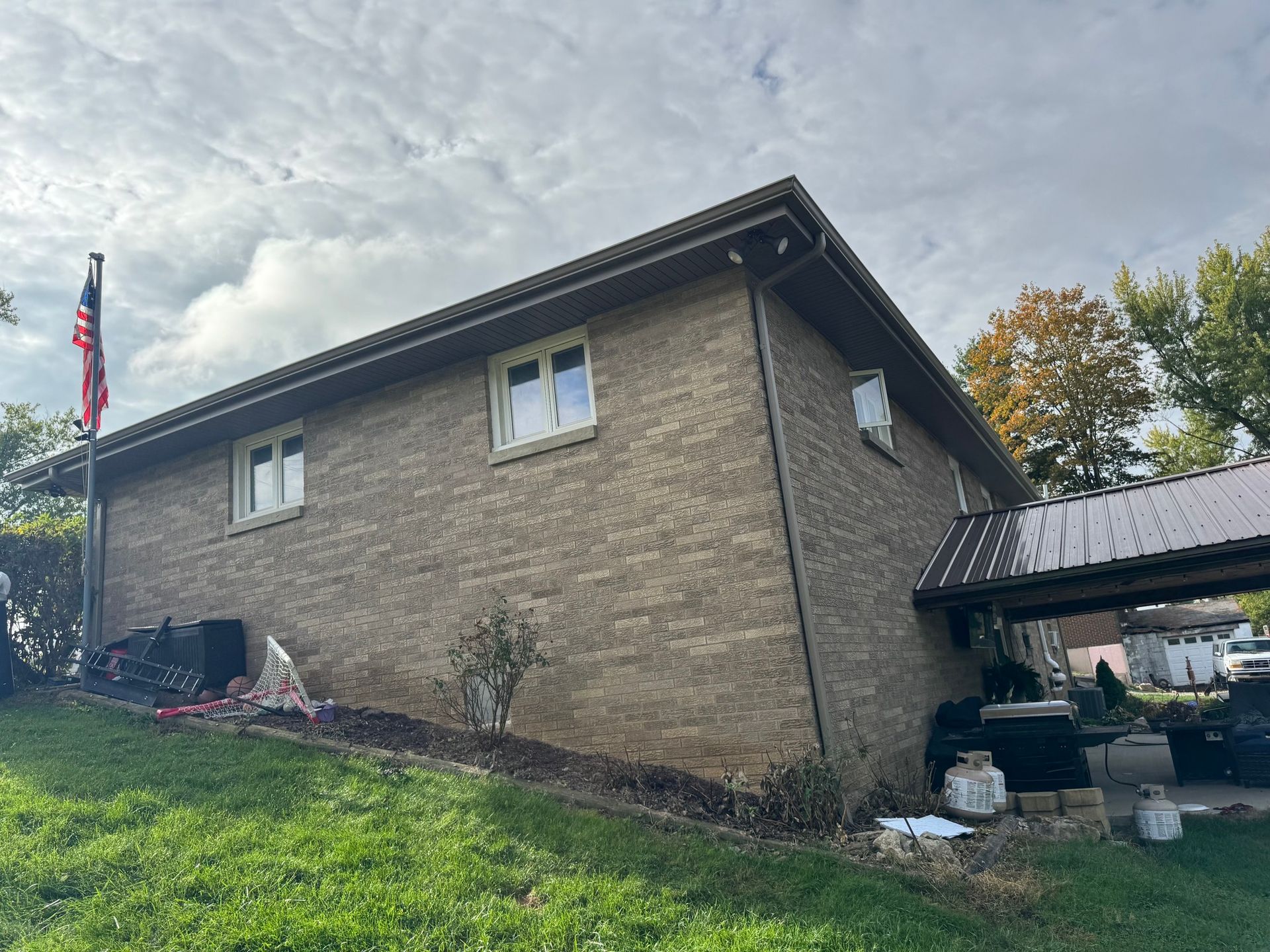 Tan brick house with two windows, a carport, and an American flag on a cloudy day.