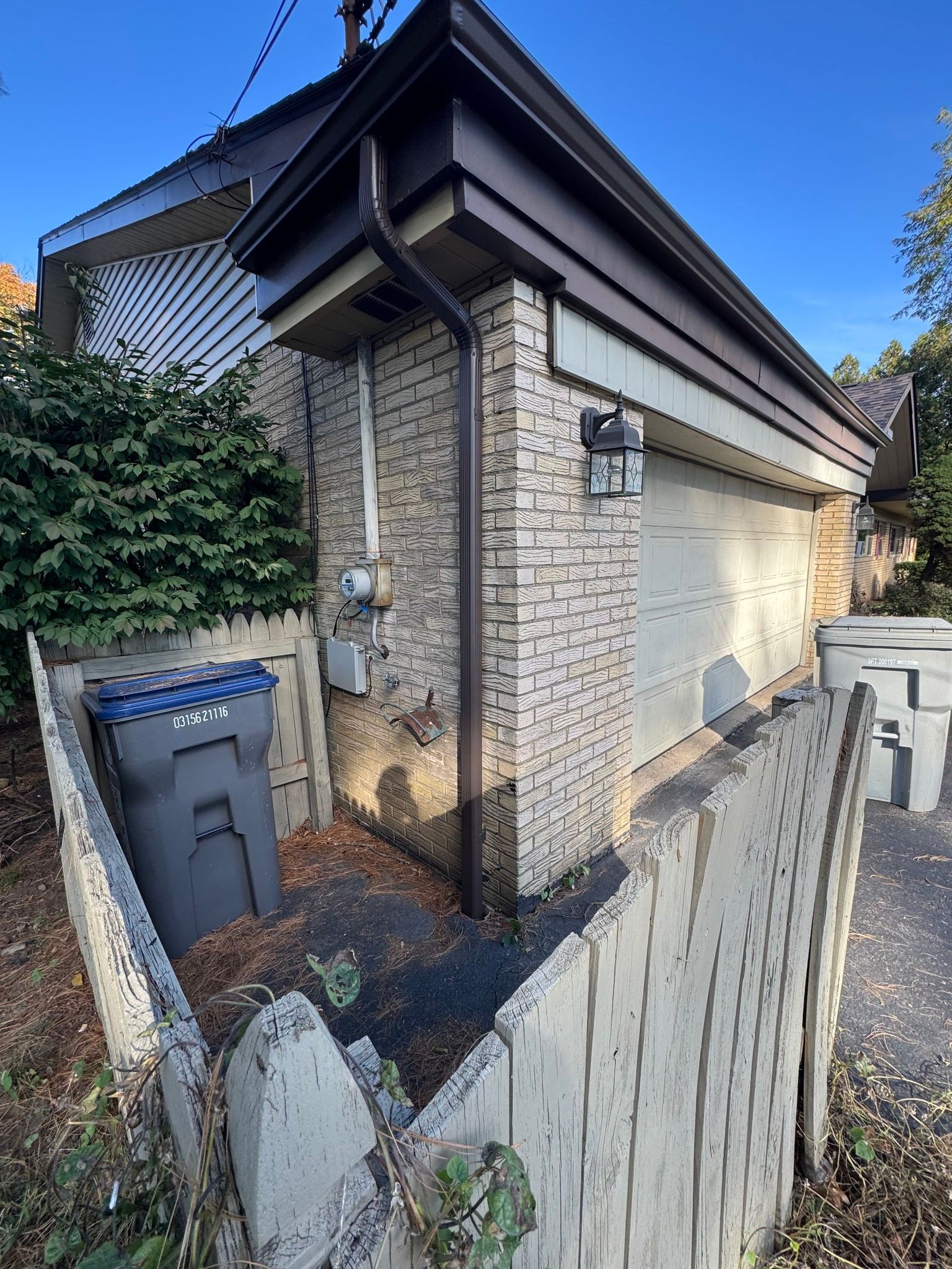 Garage with light-colored brick, white garage door, and wooden fence. A trash can sits near the garage.