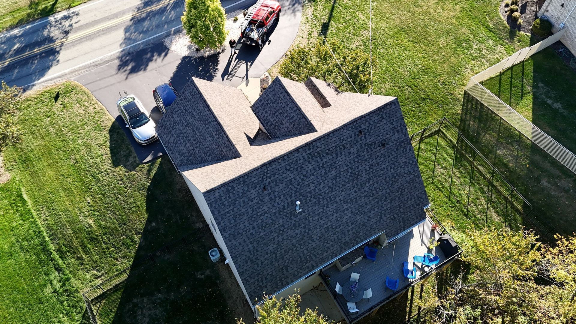 Aerial view of a house with a brown roof, driveway with cars, and a grassy yard.