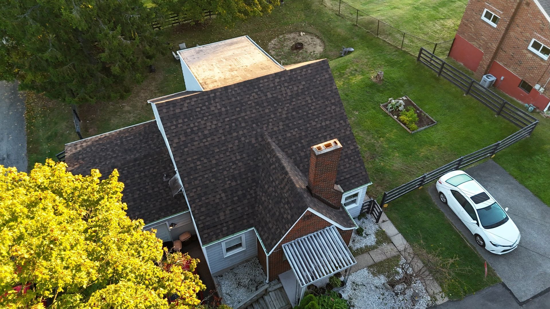 Aerial view of a house with a brown roof, chimney, and a white car parked in the driveway. Green lawn, trees, and fence.