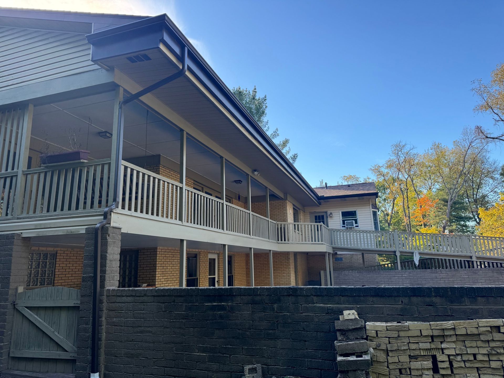 Two-story beige house with a wraparound porch, a dark roof, and a black brick wall in front.