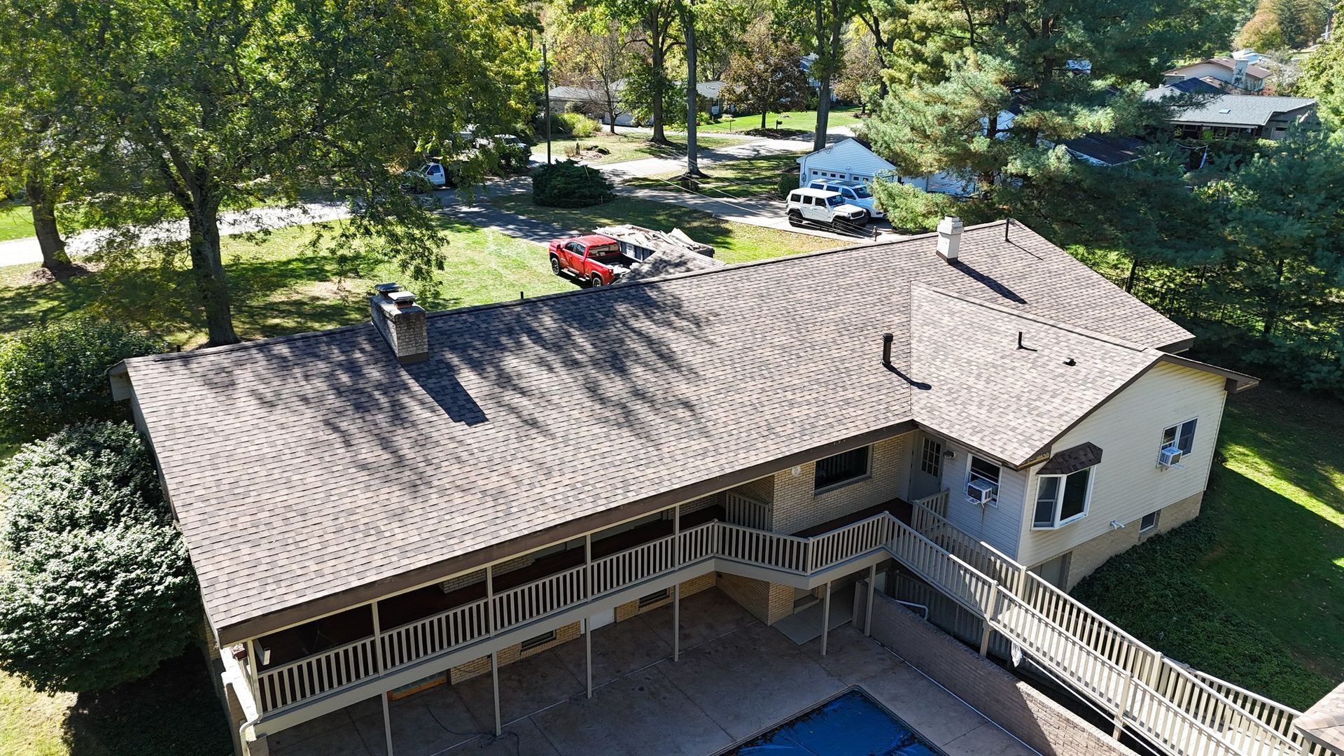 Aerial view of a house with a brown shingle roof, a pool, and surrounding green lawn and trees.