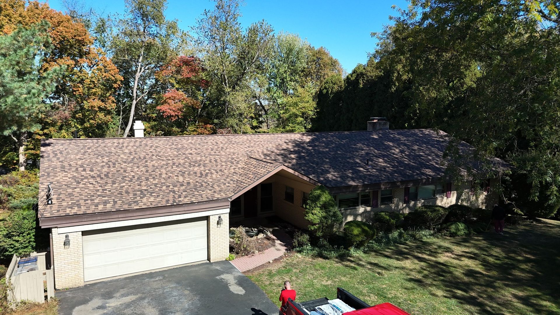 Brown-roofed house with attached garage, trees in the background, parked vehicle in driveway under a clear, blue sky.