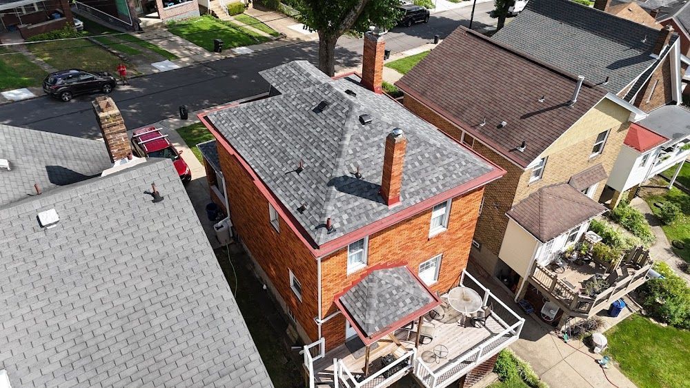 Aerial view of brick house with deck, surrounded by other houses and a street. Gray and brown roofs.