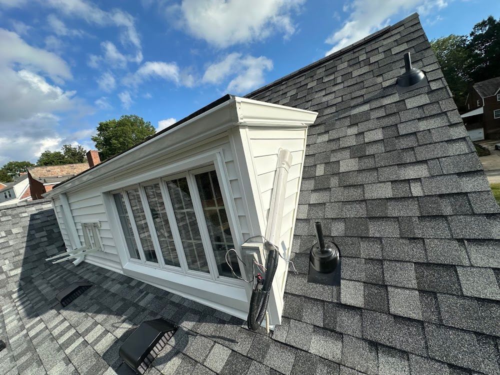 A rooftop with a white-trimmed window dormer, gray asphalt shingles, and a blue sky.