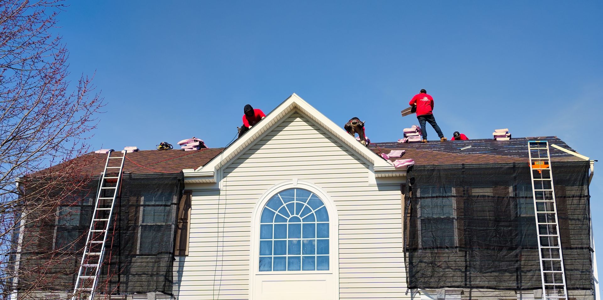 Roofers work on a house under a clear, blue sky. Ladders lean against the structure.