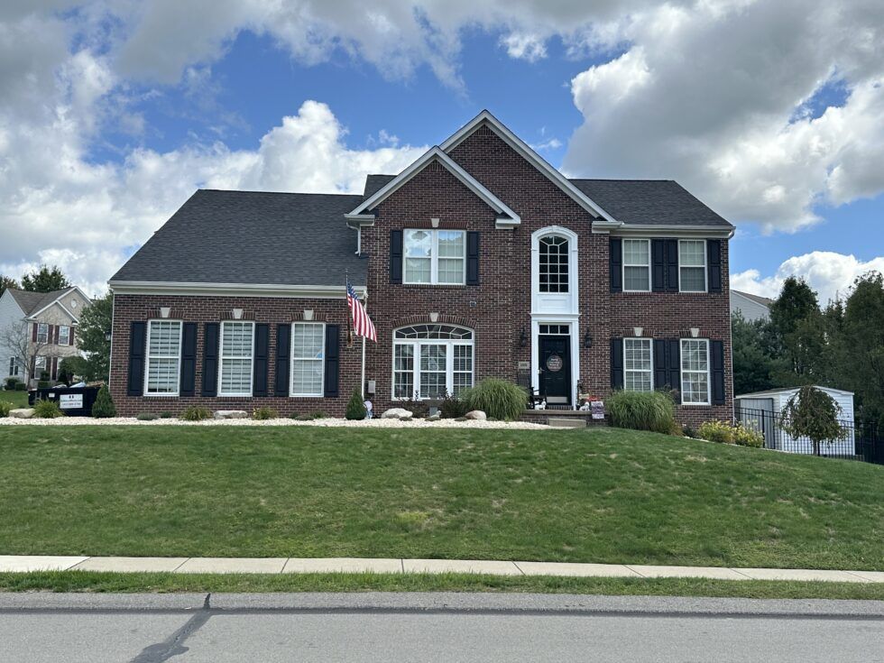 Two-story brick house with black shutters, on a grassy hill, under a partly cloudy sky.