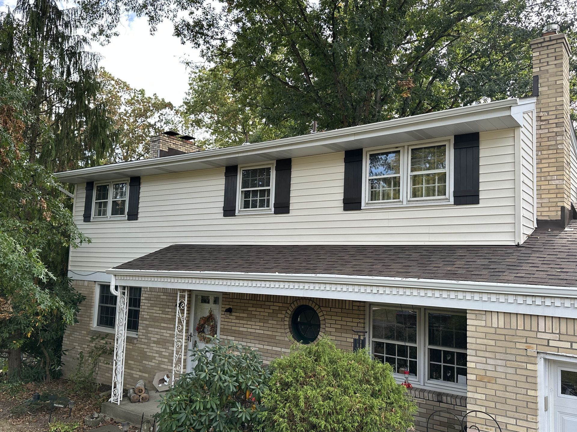 Two-story house with tan siding, black shutters, brick facade, and a chimney; trees in the background.