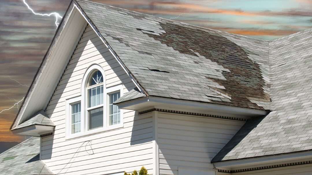 Damaged gray roof of a white house with a storm background and lightning.