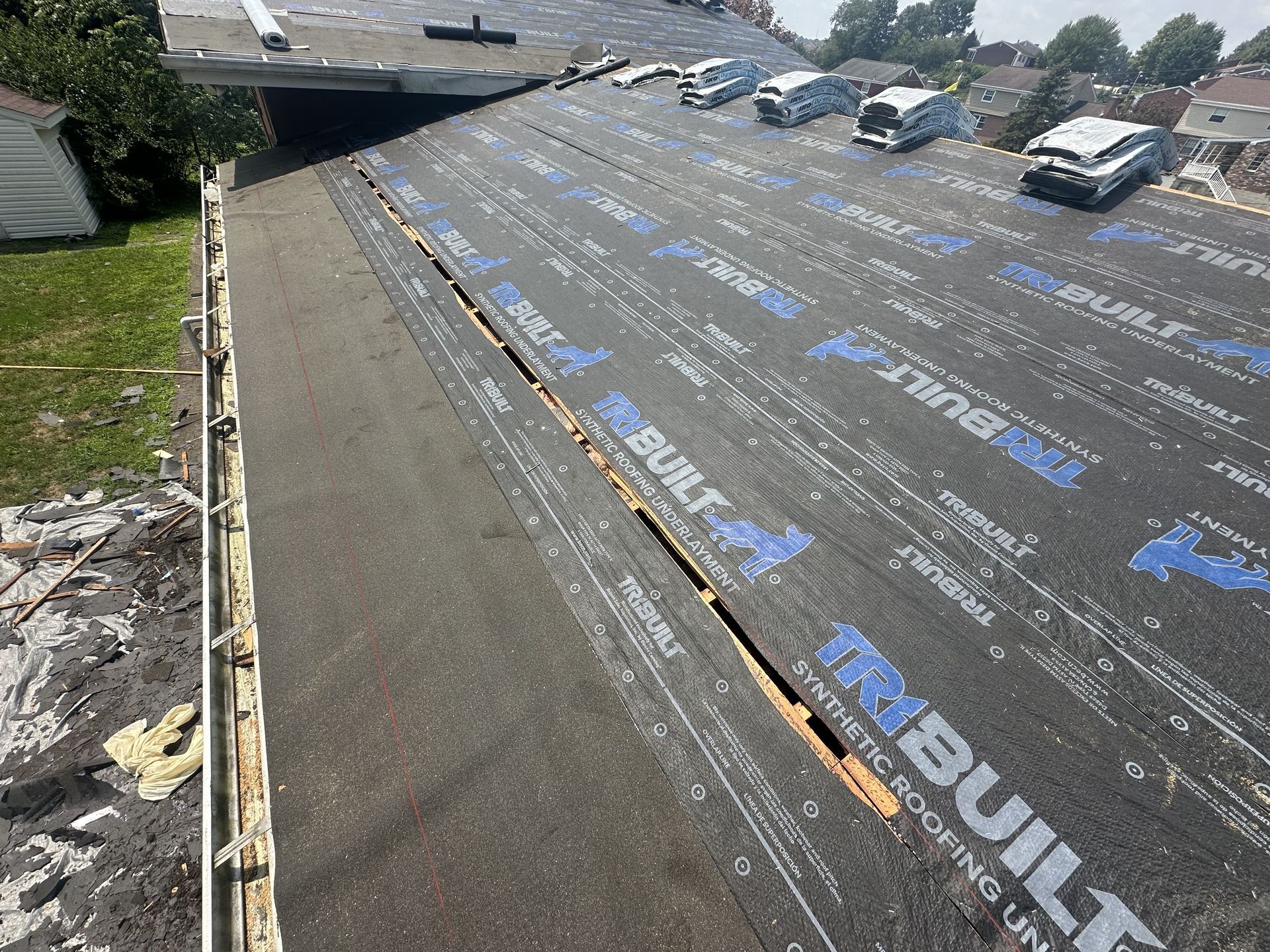 Roof under construction, black underlayment with blue branding visible, cars parked nearby.
