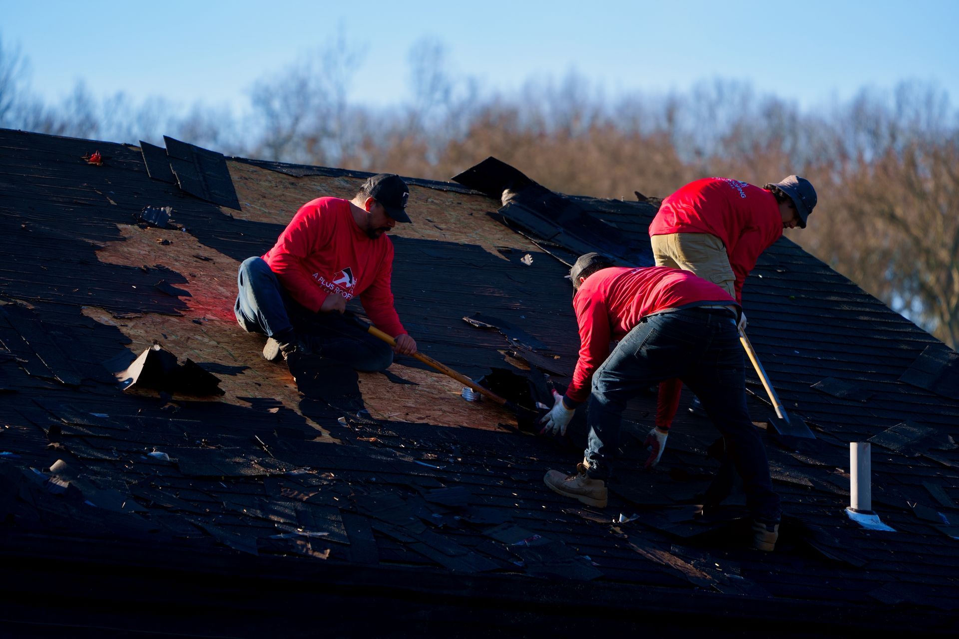 Three people in red shirts removing old roofing material from a dark roof.
