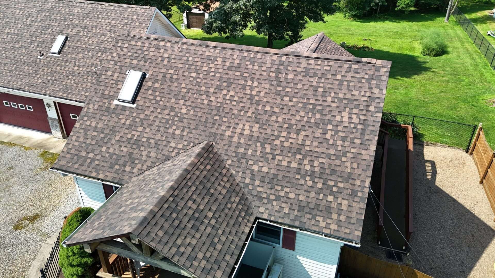 Brown shingled roof of a house with two skylights, surrounded by trees and a green lawn.