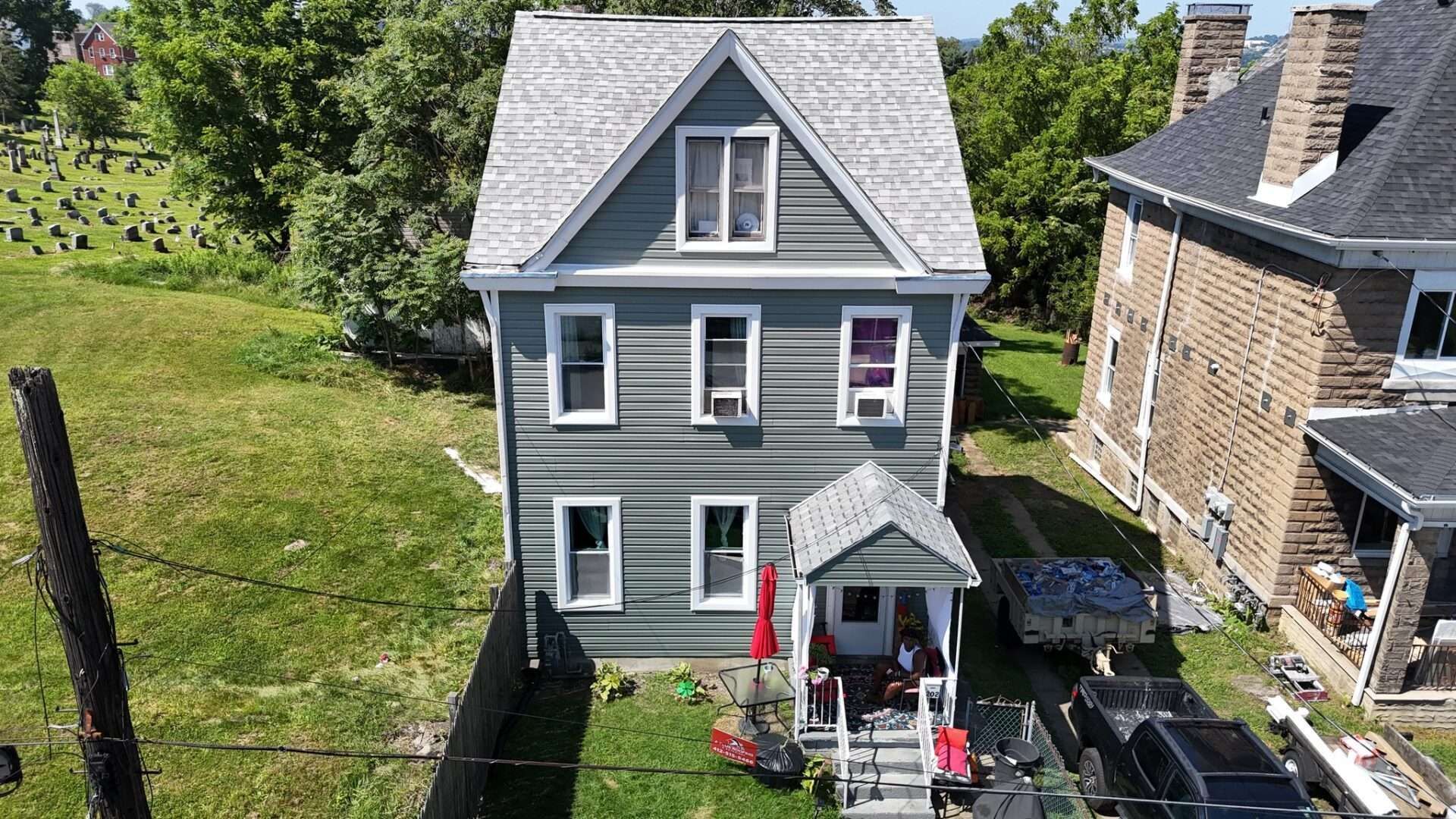 Two-story gray house with white trim. Red umbrella on porch, adjacent to a stone house.