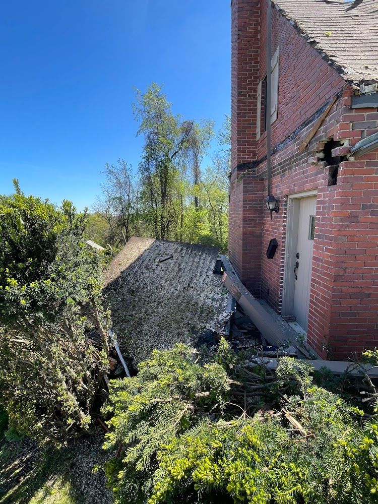 Brick building with damaged roof, surrounded by green trees and foliage under a blue sky.