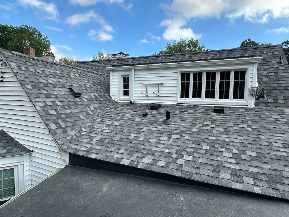 Gray shingled roof with a white dormer featuring windows, set against a partly cloudy sky.